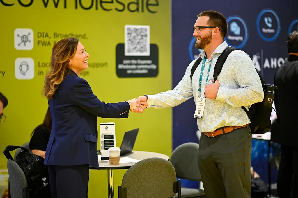 A professional networking interaction at an exhibition. A woman in a sharp dark blue suit is smiling warmly while shaking hands with a man wearing a light patterned shirt, glasses, and a backpack. In the background, a lime green display wall features corporate information, text regarding wholesale services, and a QR code.