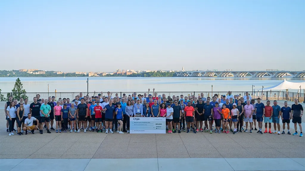 A very large group of conference attendees posing together for a group photo outdoors on a paved terrace. They are dressed in athletic running attire. In the center, a few individuals hold a large white donation check for ten thousand dollars labeled 