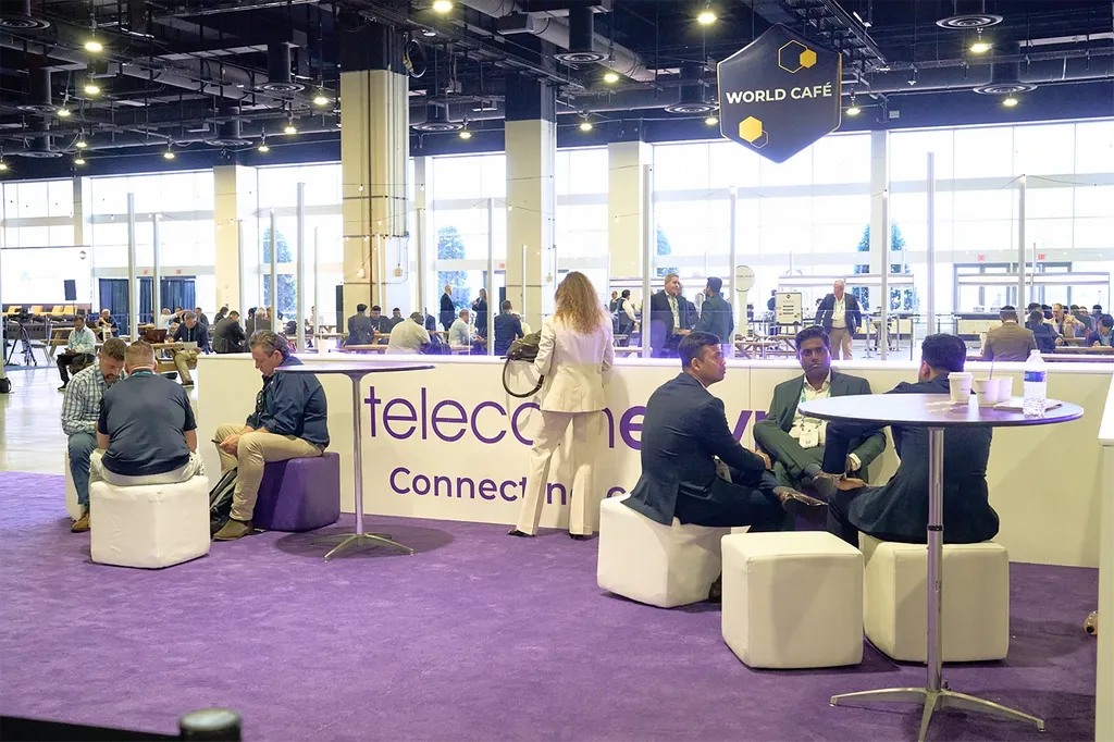 A wide shot of a bustling indoor networking lounge inside a large convention hall, set on a bright purple carpeted floor. In the foreground, business professionals are seated on white cube ottomans grouped around modern high-top tables, engaged in conversation. A large white counter in the center features purple text that reads 