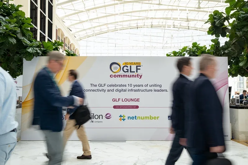 A wide, eye-level shot capturing the motion of conference attendees walking past a prominent exhibition backdrop. The attendees in the foreground are blurred to convey movement. The large white backdrop in the center displays the 