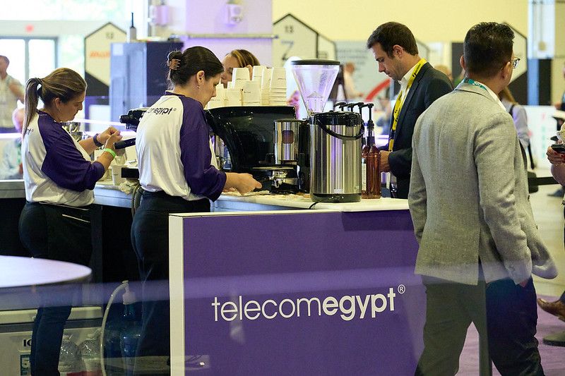 A professional event setup featuring a purple Telecom Egypt branded coffee counter. Two baristas wearing white and purple uniforms are actively working on an espresso machine and preparing drinks. Several conference attendees are standing around the counter; one man in a grey suit jacket stands in the foreground facing the barista, while others wait nearby. A large stainless steel coffee grinder and beverage dispenser sit on the counter.