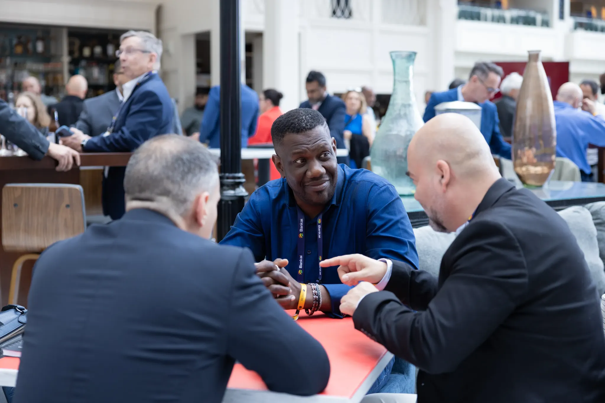 A medium shot of three business professionals having a conversation around a red-topped table at an indoor event venue. The central figure, a man in a blue button-down shirt wearing a purple 