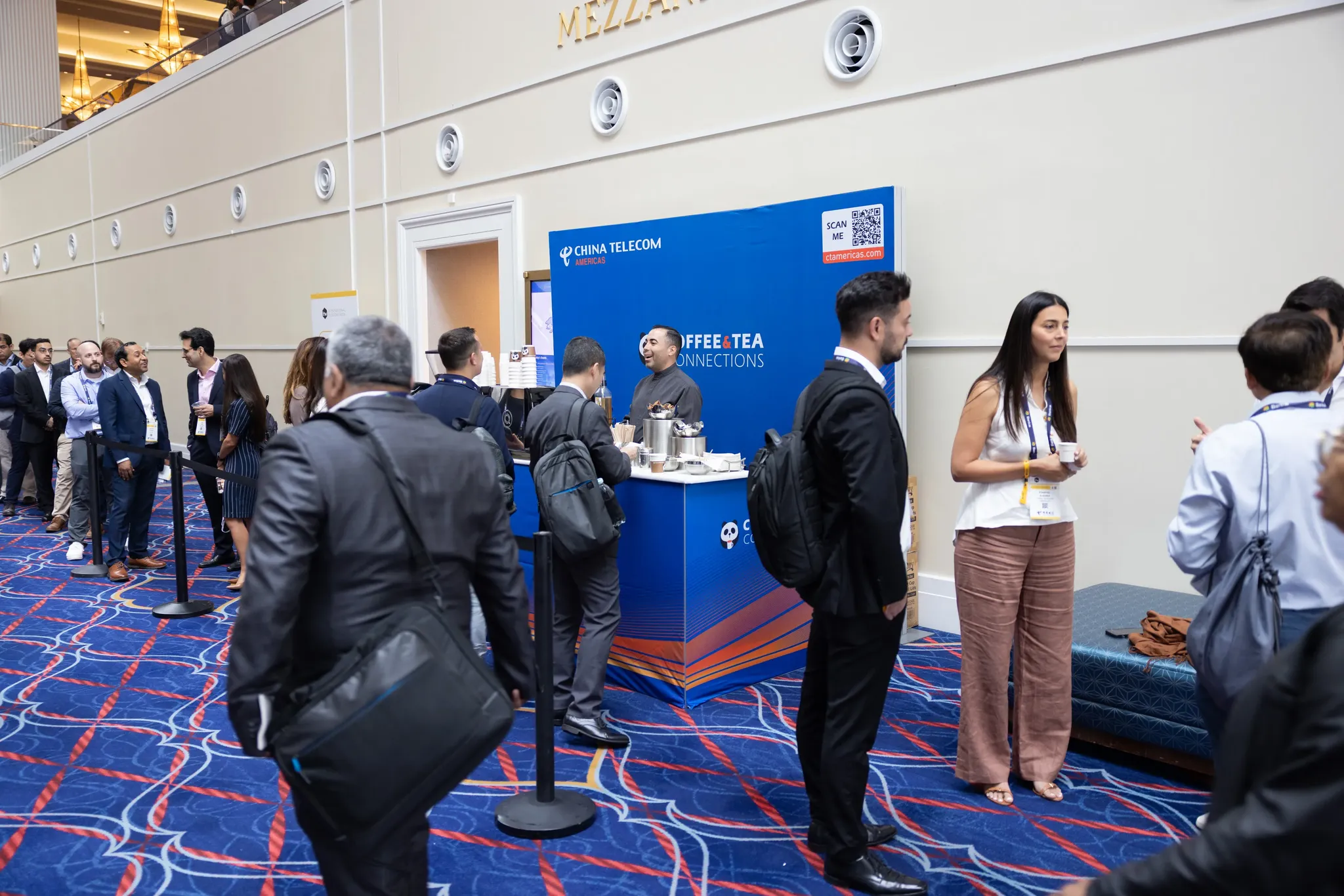 A high-angle view of a busy convention corridor with a patterned blue and orange carpet. In the center, a blue pop-up beverage stand features the 
