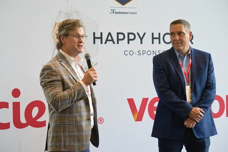 A medium shot of two men standing in front of a white exhibition banner. On the left, a man with wavy light brown hair and glasses, wearing a checkered brown blazer, holds a microphone and speaks. On the right, a man in a navy blue suit jacket with a Verizon lanyard stands with his hands clasped, listening. The background banner features the words 