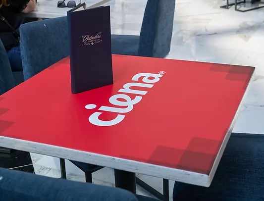 A high-angle close-up shot of a cafe-style square table at an event venue. The entire surface of the table is covered in a red vinyl wrap displaying a large white 