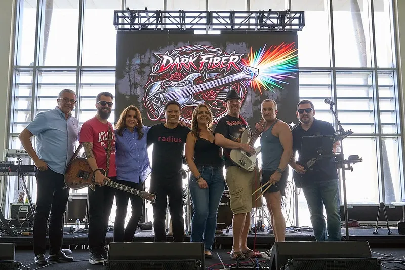 A wide, front-facing shot of eight individuals smiling and posing together on an indoor stage. Some members hold musical instruments, including an electric guitar and a bass guitar. Behind them, a massive LED screen displays a colorful 