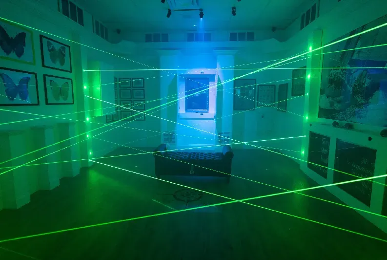A wide shot of an interactive museum or entertainment room configured as a laser maze challenge. Bright green laser beams crisscross through the dimly lit room at various angles and heights from floor to ceiling. In the center sits a dark, tufted leather bench on a wooden floor. The white walls are decorated with framed pictures of butterflies on the left and other artwork on the right, all bathed in a cool blue and green ambient glow from the overhead stage lighting.
