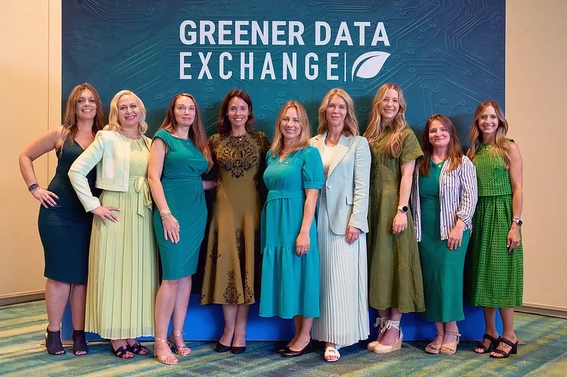 A wide, front-facing shot of nine smiling women standing in a line at a corporate event. They are dressed in a coordinated palette of greens, ranging from mint and teal to olive and forest green—along with patterned earth tones. They are posing in front of a large, dark teal backdrop with circuit board patterns that reads 