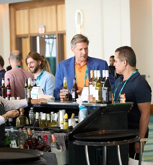 A medium shot of a social gathering around a bar. In the center, a man in a vibrant blue blazer over an orange polo shirt holds a beer bottle while conversing with a man in a dark polo shirt. The bar counter in the foreground is lined with wine, beer, and liquor bottles, with a bartender's hands visible on the left. In the background, other attendees wearing conference lanyards are seen smiling and talking in a brightly lit, modern venue.