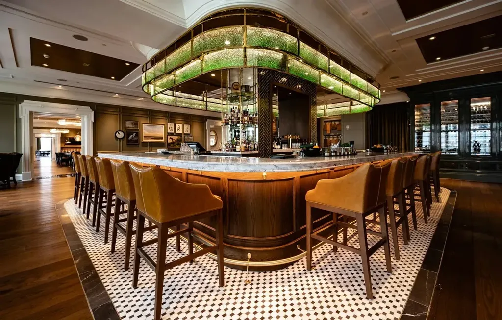 A wide shot of a luxurious indoor bar area. The central, curved wooden bar has a white marble countertop lined with a row of high-backed, tan leather barstools with wooden legs. Hanging directly above the bar is a massive, oval-shaped light canopy with brass trim and a textured green glass design that glows from within. The bar area is set on a black-and-white checkered tile floor bordered by dark hardwood flooring. In the background, muted olive-green walls feature framed artwork, and an ornate dark wood wine cabinet stands on the right.