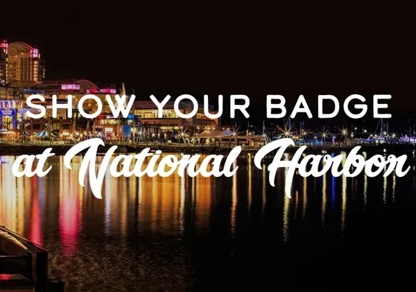 A promotional event graphic featuring a night scene at National Harbor. Brightly lit commercial buildings, restaurants, and a marina area line the background, casting vibrant orange, yellow, and red light reflections across the dark water in the foreground. Large text is overlaid across the center of the image, reading 