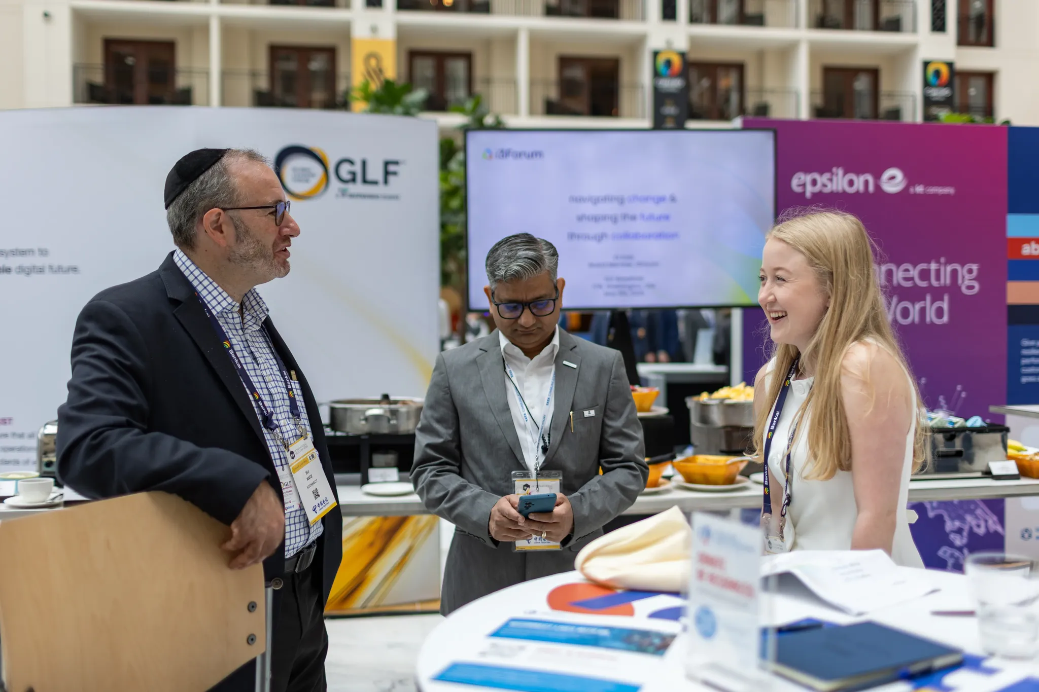 An indoor photograph showing three attendees interacting in a bright conference hall. On the left, a man wearing glasses, a patterned shirt, a dark blazer, and a black yarmulke leans against a wooden chair while speaking. On the right, a blonde woman wearing a white sleeveless top smiles warmly during their conversation. In the center, a man in a grey suit and glasses looks down at his smartphone. The background features exhibition banners with 