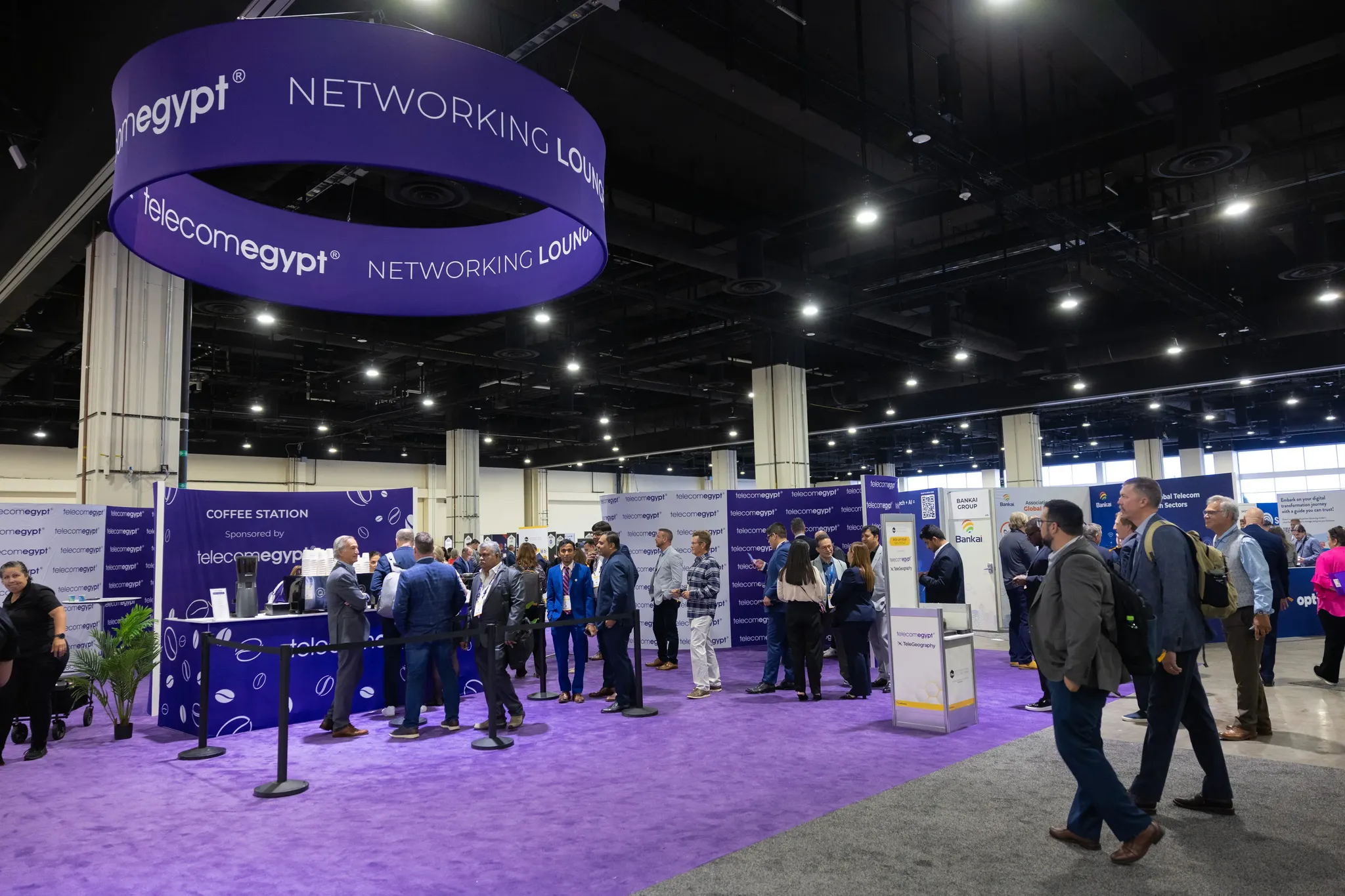 A wide shot of a spacious corporate convention hall. A designated networking area features vibrant purple carpeting and a massive, circular purple banner hanging from the ceiling that reads 