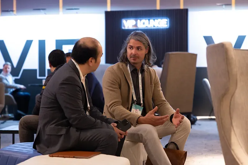 A medium shot of two men conversing at an event. On the left, a man with thinning hair sits in profile wearing a dark grey blazer, holding a phone in his hands. On the right, a man with long, wavy greyish-brown hair wears a beige blazer and light trousers, gesturing with both hands as he speaks. They are in a modern lounge area with a glowing blue 