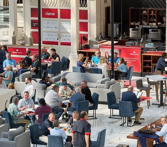 A high-angle, vertical shot of an open-air indoor pavilion or courtyard converted into a corporate networking area. Numerous conference delegates are seated at dining tables, high-top bars, and grey modern lounge couches scattered across a marble-tiled floor. In the background, a white architectural structure features a large red wall banner reading 