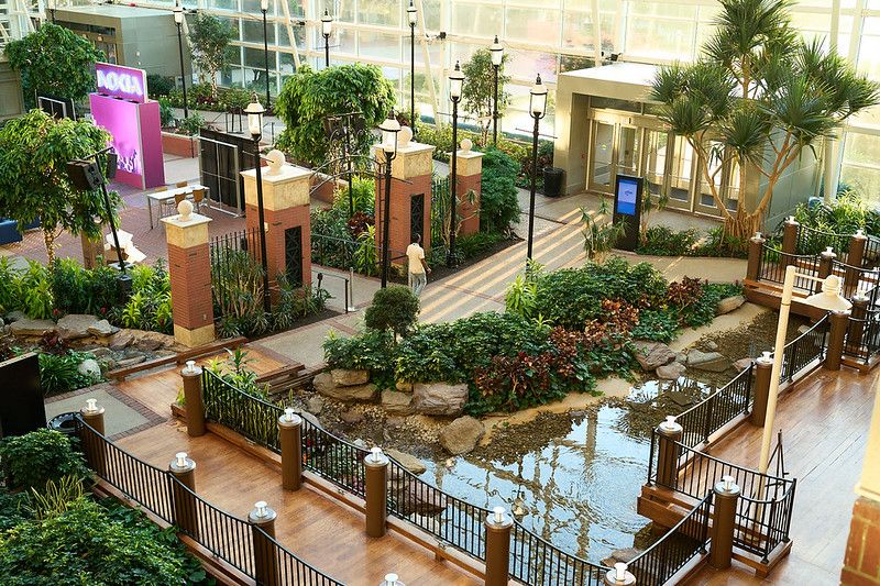 A high-angle, wide shot of a lush indoor atrium featuring a landscaped garden and a water feature. Wooden walkways and bridges with dark metal railings cross over a rocky, shallow pool of water. The space is filled with dense green bushes, small trees, tropical plants, and classic black lampposts. In the upper left background, a pink event installation with a glowing Nokia logo is visible near brick pillar gates, all housed under a massive glass ceiling that floods the area with natural light.