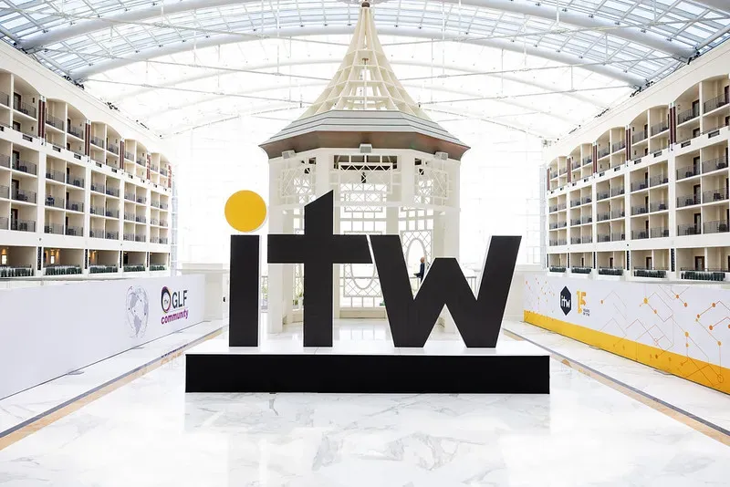 A wide, straight-on shot of a large corporate event setup inside a bright, white resort atrium. In the center, a large, free-standing black sculpture displays the lowercase letters 
