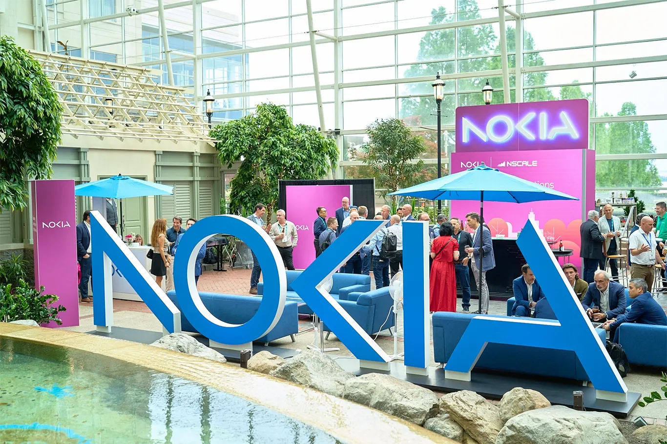 A wide shot of a brightly lit indoor atrium serving as a Nokia-branded networking lounge at a convention. In the foreground, a large, free-standing blue sculpture spells out the word 