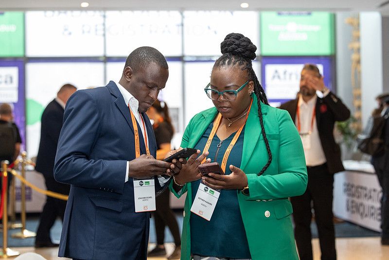 Two conference attendees wearing event badges use their mobile phones in the exhibition and registration area of a business conference.