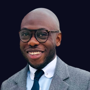 Headshot of Jacques Abeng wearing dark-rimmed glasses, a grey textured coat over a light blue collared shirt, and a dark navy blue tie. He is set against a solid dark background.