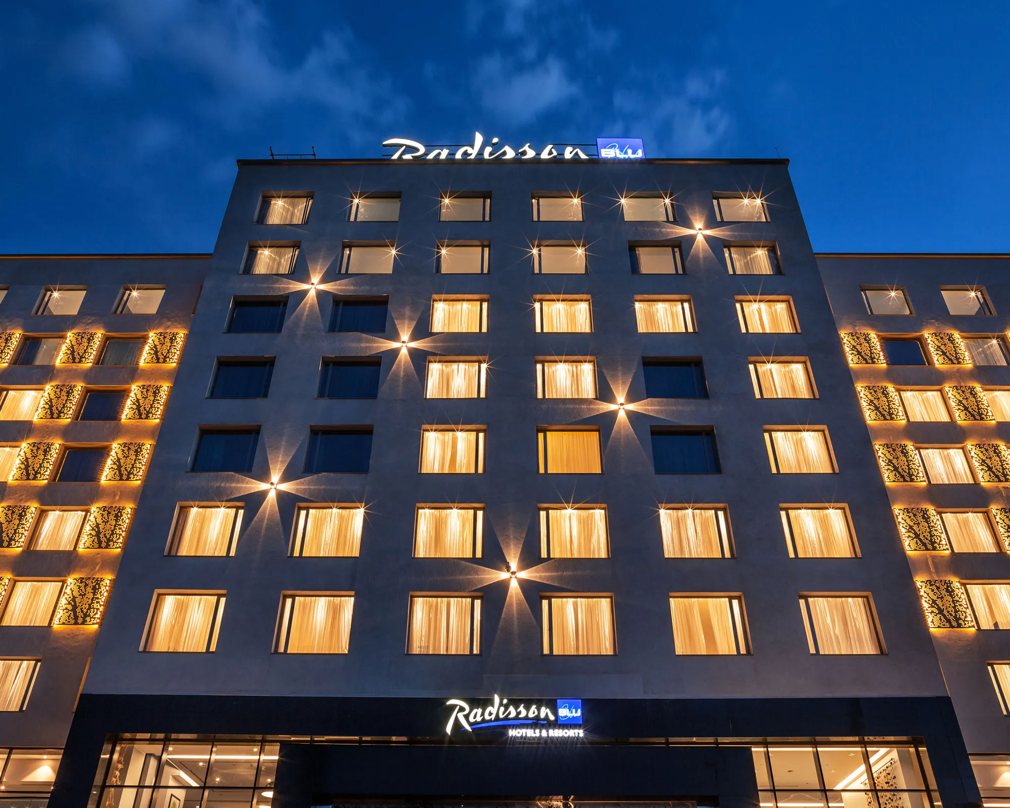 A low-angle, evening view of the modern multi-story facade of the Radisson Blu Hotel. The building's grid of windows is warmly illuminated from within, while exterior accent lights create sharp starburst patterns across the structure. Decorative, glowing golden panels flank the sides of the central concrete building. The illuminated 