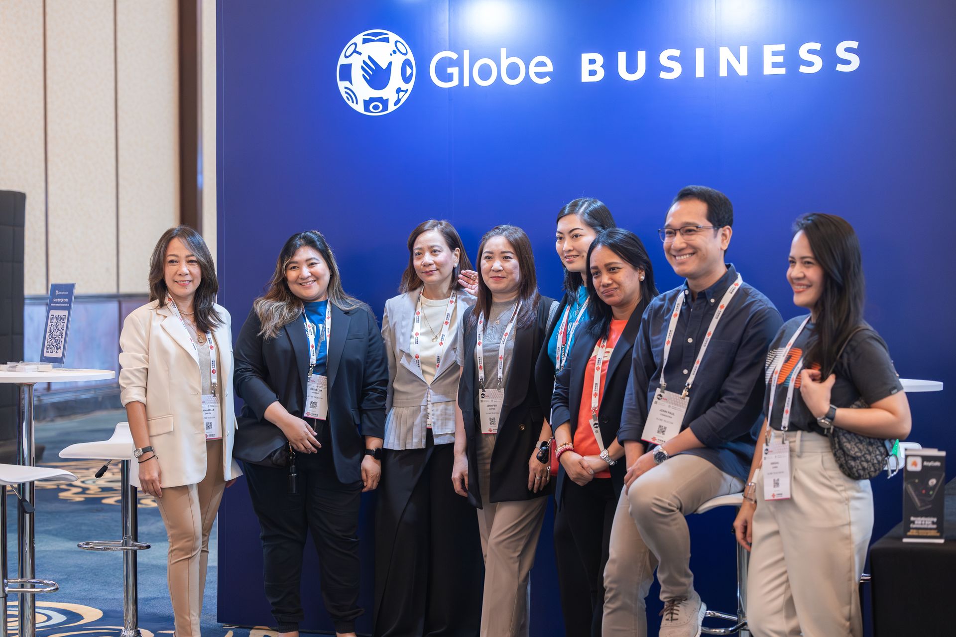 A group of eight business professionals posing for a picture at a corporate convention. They are standing or leaning on bar stools in front of a solid blue exhibition wall that displays the white 