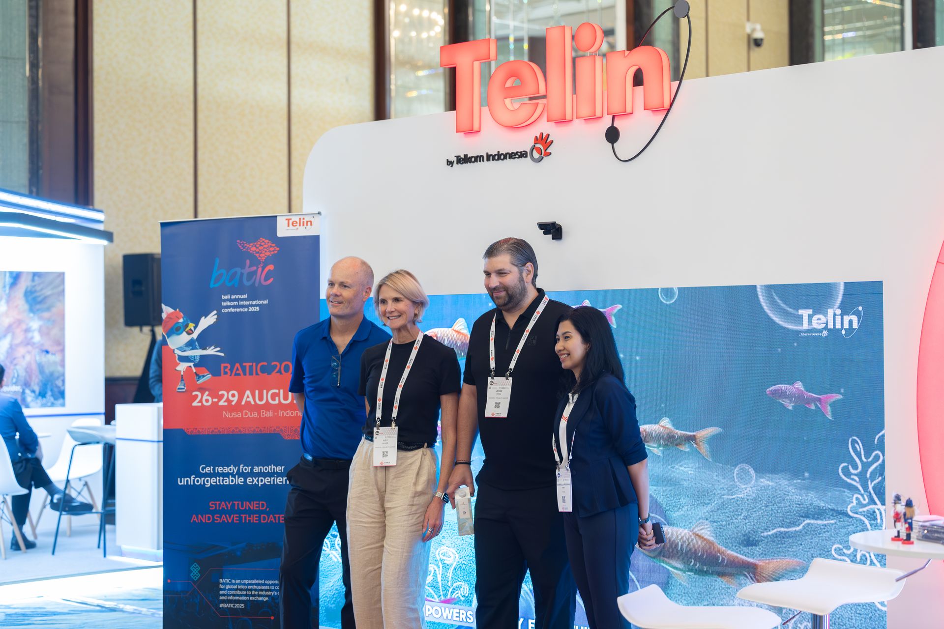 Four corporate professionals stand posing for a photo together at an indoor exhibition booth. The white backdrop wall features a prominent red illuminated sign reading 