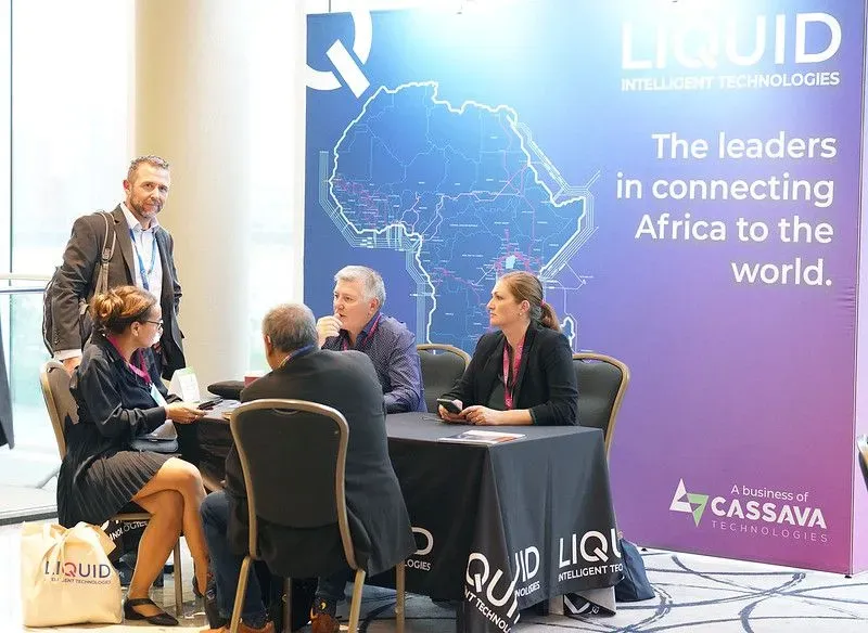 A photograph of a networking area or exhibition booth for Liquid Intelligent Technologies at a conference event. Five people are gathered around a small table covered with a branded black tablecloth; four are seated in discussion, while one man in a suit stands to the left looking toward the camera. A large purple and blue backdrop behind them displays a network map of Africa, the company logo, and the text 