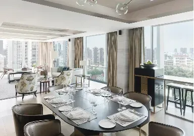 A photograph of a bright, modern open-plan living and dining area with large floor-to-ceiling windows overlooking a city skyline. In the foreground, a dark oval dining table is fully set with placemats, plates, silverware, and glassware, surrounded by dark upholstered chairs. The background features a lounge area with a light-colored sofa, two floral-patterned armchairs, and a coffee table. On the right side, there is a dark modern cabinet next to a window-facing counter with two bar stools. Clear spherical pendant lights hang from the ceiling above.