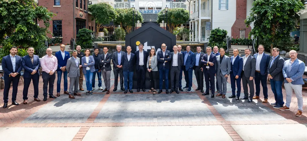 A group of diverse business professionals posing together outdoors in a paved courtyard. The group is standing in front of a large, black monument featuring the 