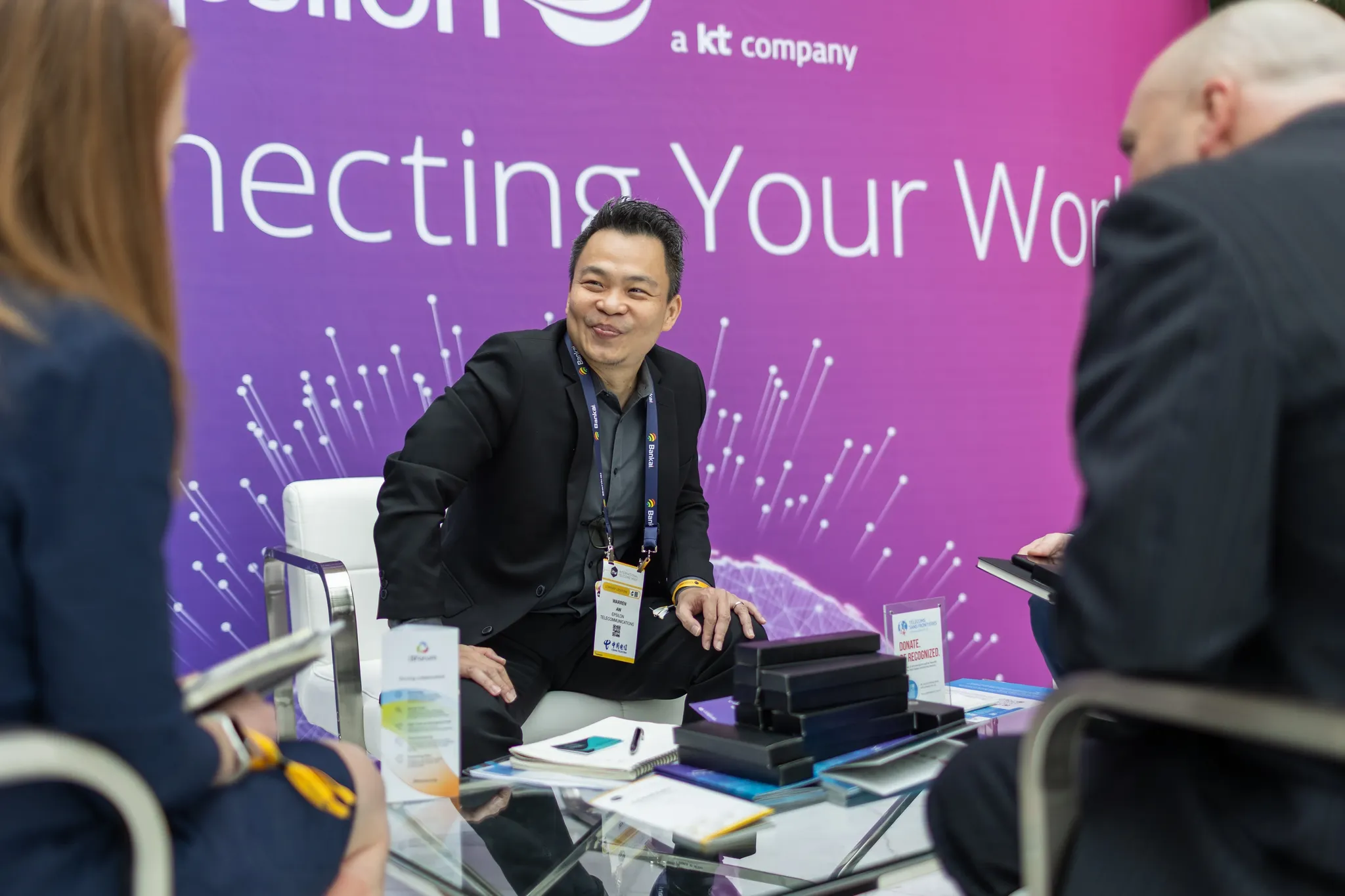 A professional networking scene at a GLF event. A man in a black suit and lanyard sits in a white chair, smiling and engaging in conversation with two colleagues at a glass table. In the background, a large purple banner features the phrase 