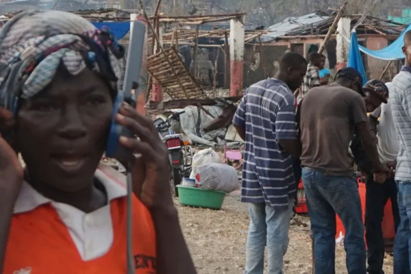A close-up of a person wearing a headwrap and an orange shirt, speaking into a large satellite phone. The background shows a crowded area with damaged structures and several people standing near what appears to be a makeshift camp or disaster relief site.