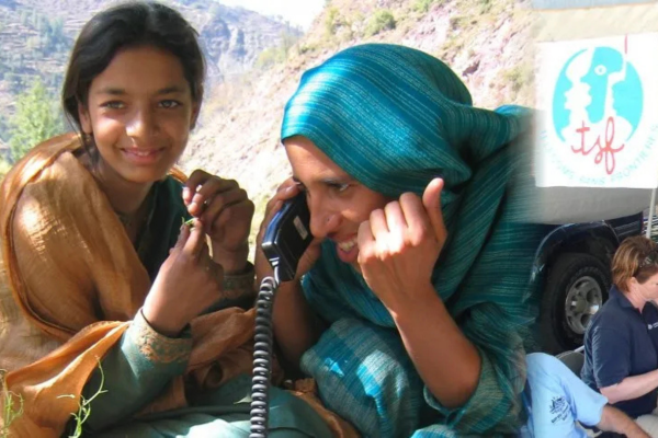 Two people sitting outdoors against a mountainous backdrop; one smiles at the camera while the other speaks on a corded handset. On the right, a logo for T&eacute;l&eacute;coms Sans Fronti&egrave;res (TSF) is visible on a white banner next to relief workers and a vehicle.