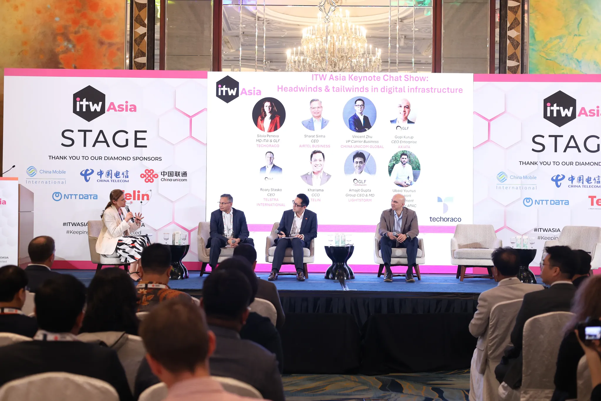 A photograph of a panel discussion taking place on stage at the ITW Asia conference. Four panelists—one woman and three men—are seated in light-colored armchairs holding microphones, facing an audience whose heads and shoulders are visible in the foreground. Behind the speakers, a large central screen displays the text 