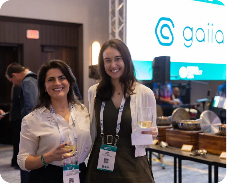 Two smiling women stand side-by-side posing for a photo at a professional conference. Both wear conference lanyards around their necks and hold glasses of white wine. In the background, other attendees, a buffet station, and a large digital screen with the 