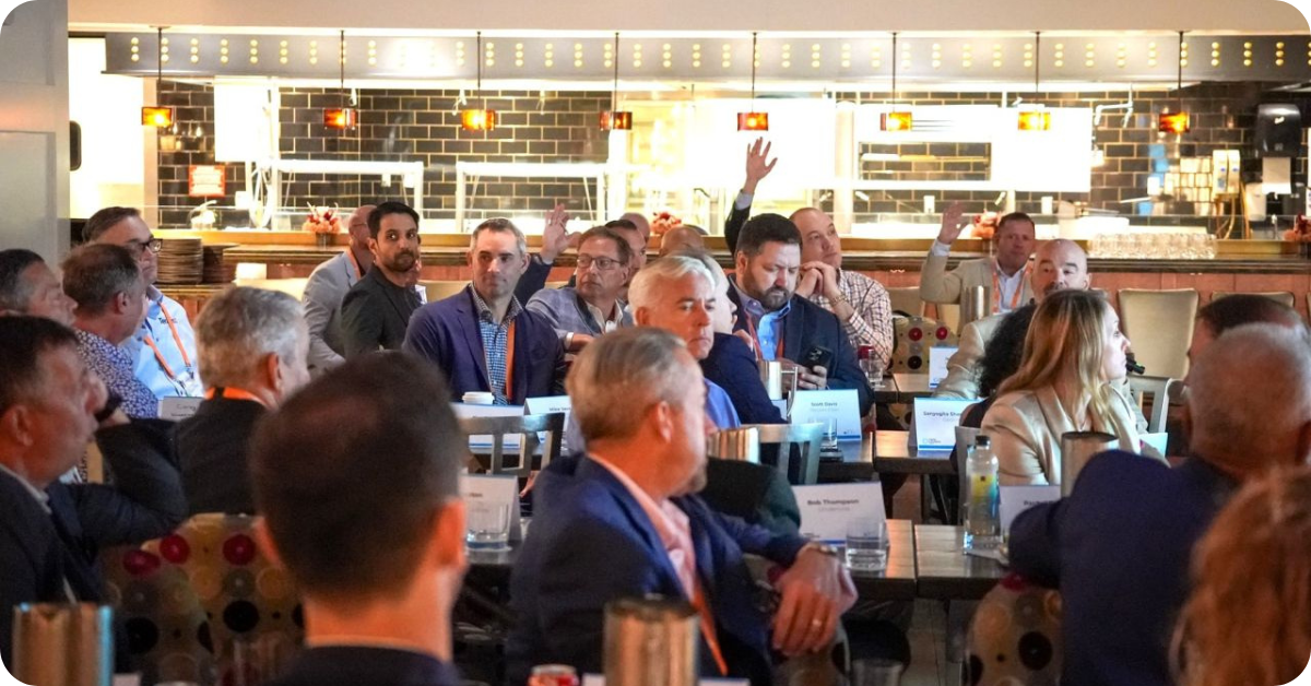 Fiber leaders seated around tables during a group discussion at the Fiber Leaders Lunch, with several attendees raising their hands to contribute.