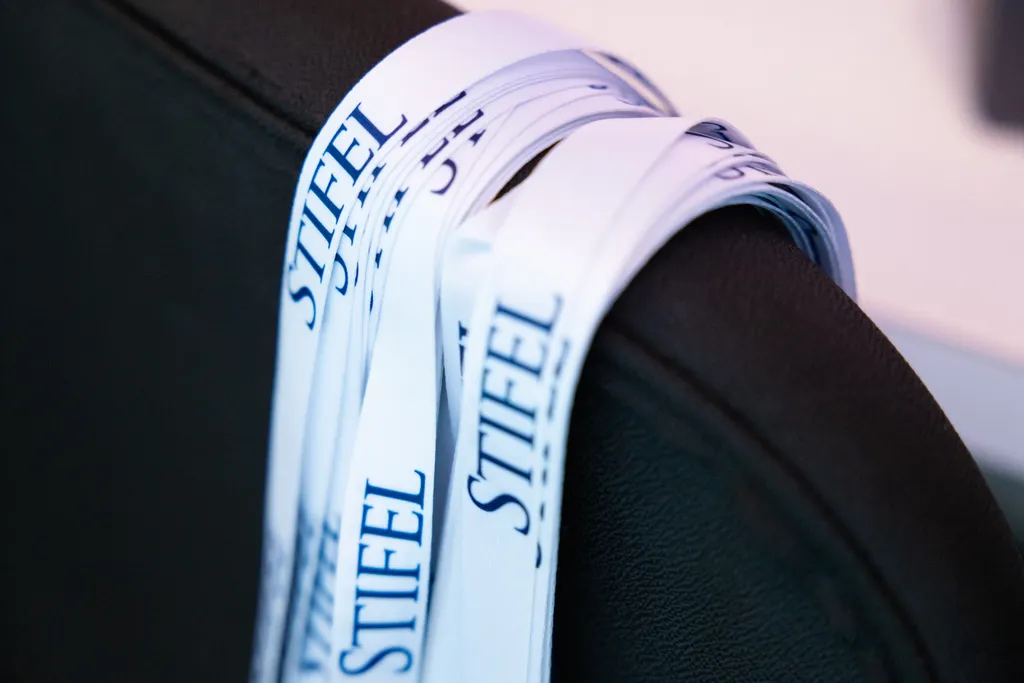 A close-up, shallow-depth-of-field shot of several white fabric event lanyards looped over the top curve of a dark grey or black office chair. The lanyards feature the corporate name 
