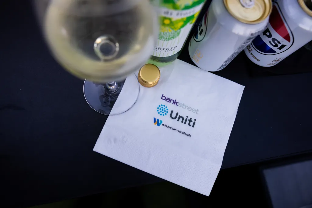 A top-down, close-up shot of a white cocktail napkin resting on a dark tablecloth. The napkin features three printed corporate logos stacked vertically: Bank Street, Uniti, and Windstream Wholesale. Surrounding the napkin are a partially visible glass of white wine, a green wine bottle neck with a gold screw cap, a can of Diet Pepsi, and another white soda can.