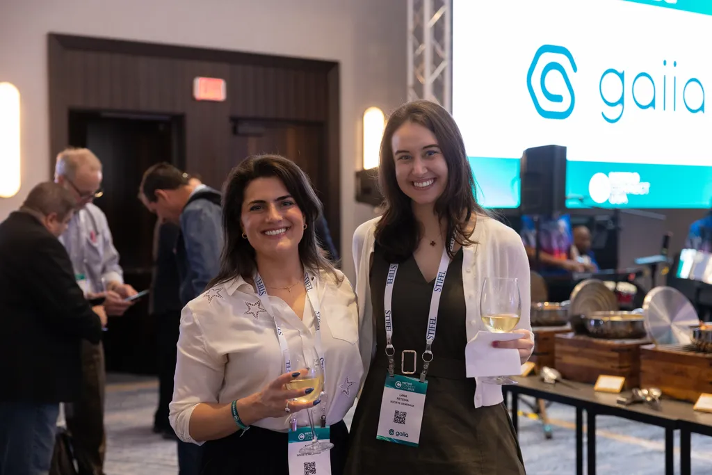 Two smiling women stand side-by-side posing for a photo at a professional conference. Both wear conference lanyards around their necks and hold glasses of white wine. In the background, other attendees, a buffet station, and a large digital screen with the 