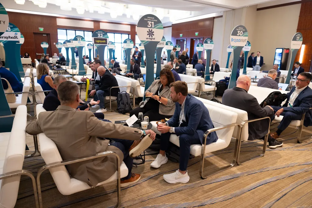 A high-angle, wide shot of a bustling corporate networking lounge inside a large hall. Professionals are seated in pairs and small groups on modern white leather armchairs arranged face-to-face. Tall, teal, surfboard-shaped signs topped with round lounge numbers and sponsor logos (such as 