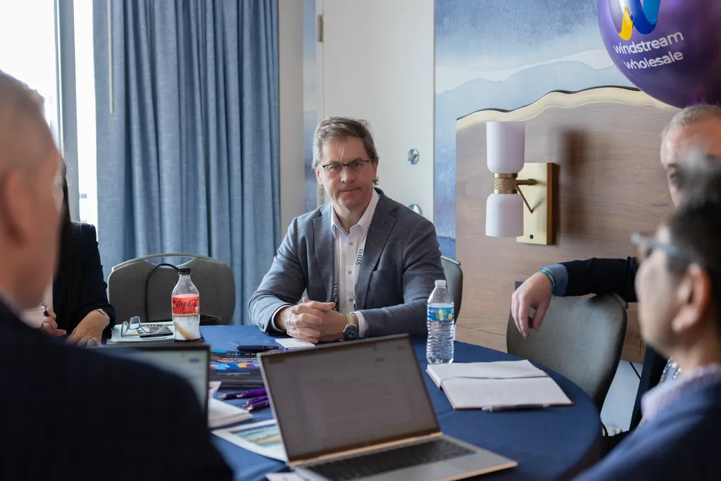 A group of professionals sitting around a blue fabric-covered circular table during a business meeting. A man in a grey blazer and glasses sits at the center with his hands clasped, listening. Laptops, notebooks, water bottles, and a purple 
