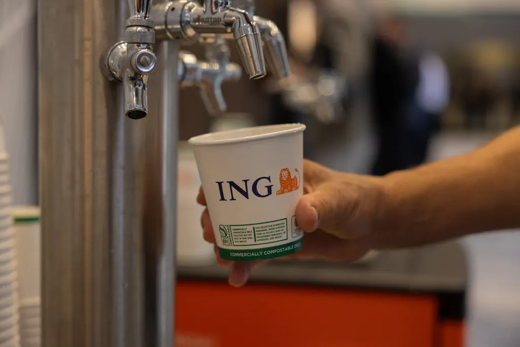 A tight, shallow-depth-of-field shot of a person’s hand holding a white paper cup under a shiny stainless steel draft tap faucet. The cup prominently features the orange lion logo and blue text for the financial company ING, along with text stating 