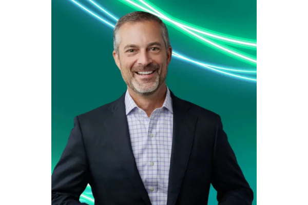 Professional headshot of Chad Crank with short grey-flecked hair and a trimmed beard, wearing a dark suit jacket over a light purple and white checkered button-down shirt. The background is teal with glowing green and white abstract lines curving across the top.