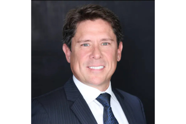 Professional headshot of Hunter Newby with short dark hair, wearing a dark pinstripe suit, a white collaired shirt, and a dark patterned tie. The background is a solid black gradient.