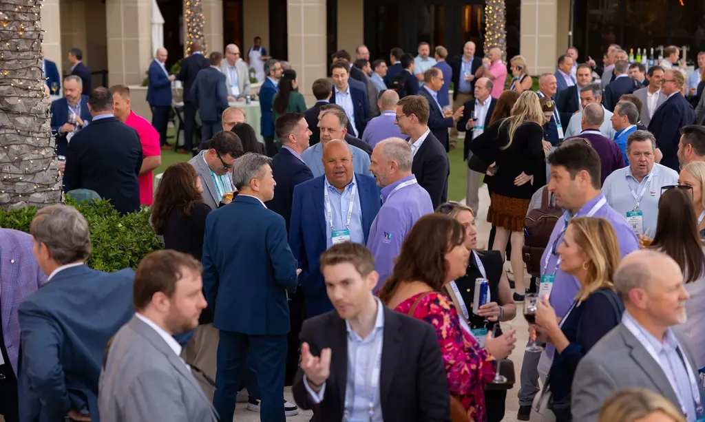 A high-angle, wide shot of a crowded outdoor networking reception at a professional conference. Dozens of attendees in business casual attire are gathered on a patio, talking in small groups while holding drinks. Palm trees wrapped in string lights and building structures are visible in the background.