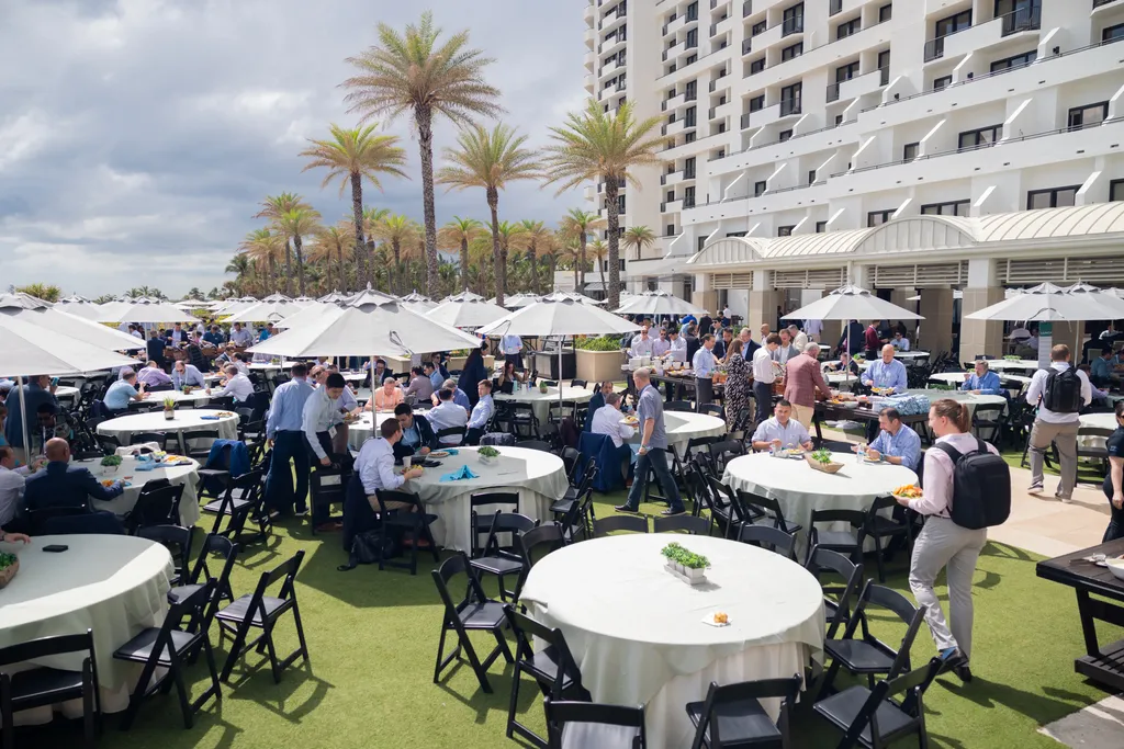 A high-angle, wide shot of a crowded outdoor luncheon at a coastal resort or conference venue. Numerous round dining tables covered in light-colored tablecloths and shaded by large white umbrellas are spread across a green artificial lawn. Dozens of professionals are seen eating, talking, or carrying plates from a buffet line setup next to a large, multi-story white hotel building. Palm trees stand in the background under a cloudy sky.