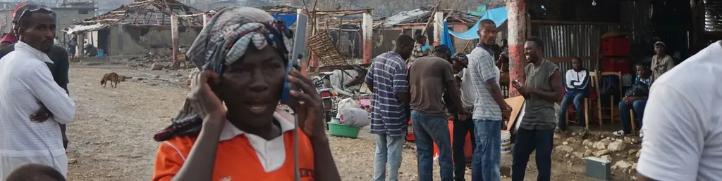 A wide outdoor shot following a disaster or crisis event, showing a bustling community area with heavily damaged, roofless wooden and concrete structures in the background. In the foreground, a woman wearing an orange collared shirt and a patterned headwrap holds a large satellite telephone to her ear, speaking intently. In the midground, a group of men stand around chatting near makeshift market stalls, while a stray dog walks through the unpaved, dusty street on the left.