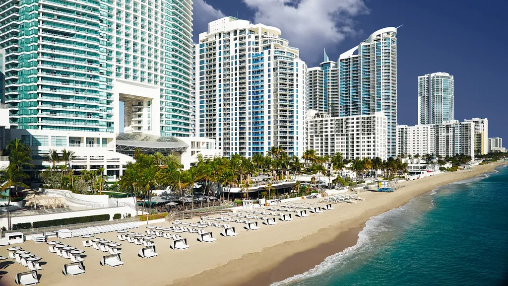 A high-angle, wide drone shot of a massive luxury beachfront resort complex. The left side is dominated by towering white and teal high-rise hotel buildings. At the base of the towers, a wide sandy beach is lined with rows of white canopy daybeds and lounge chairs, bordering a clear turquoise ocean on the right.