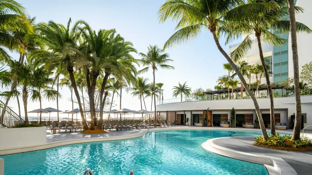 A bright, sunny view of a curved, turquoise resort swimming pool surrounded by lush tropical palm trees. In the midground, a paved patio features rows of grey lounge chairs shaded by large umbrellas, while a white multi-story hotel structure rises on the right.
