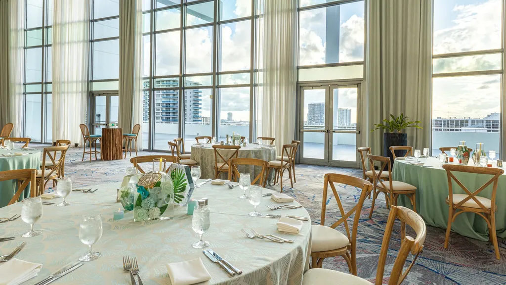 A bright, wide shot of an event setup inside a spacious hall with soaring ceilings. Massive floor-to-ceiling glass windows form the back wall, letting in abundant natural light and revealing high-rise buildings in the distance. The room features multiple round banquet tables with patterned sage-green and blue tablecloths, elegant wooden cross-back chairs, formal table settings, and floral centerpieces on a patterned carpet.